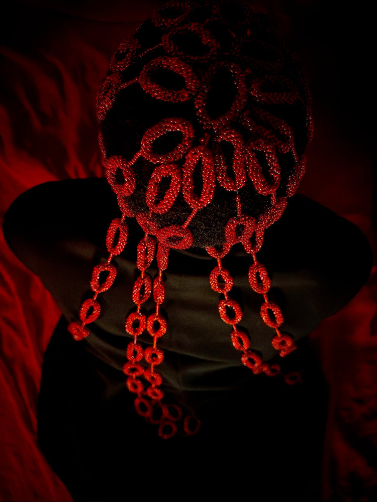 Person seated against a deep red backdrop wearing an intricate red beaded headpiece that covers the head and drapes in loops over the face and chest. Low lighting highlights the glow and texture of the beads against a dark silhouette.